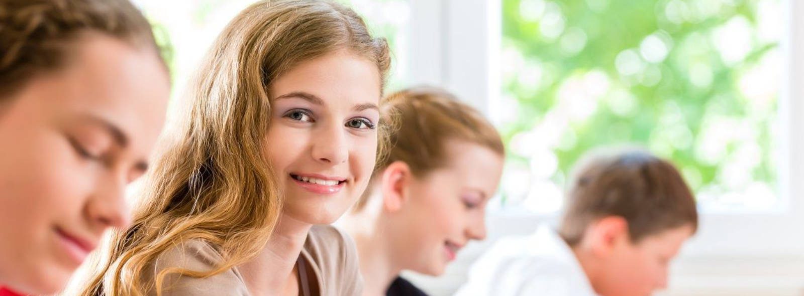 Vier Schülerinnen und Schüler sitzen nebeneinander an Tischen und schreiben, eine Schülerin blickt lächelnd in die Kamera